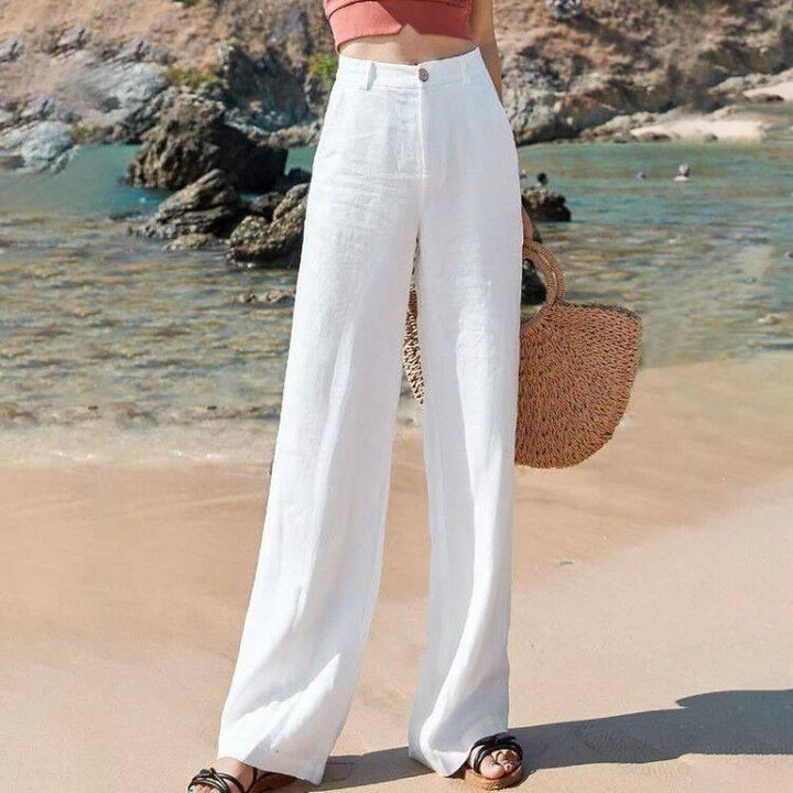 Woman wearing white wide-leg pants and a crop top on a sandy beach holding a woven straw bag