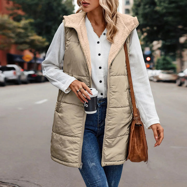 Woman in beige quilted vest with sherpa lining, white button shirt, and blue jeans holding coffee cup