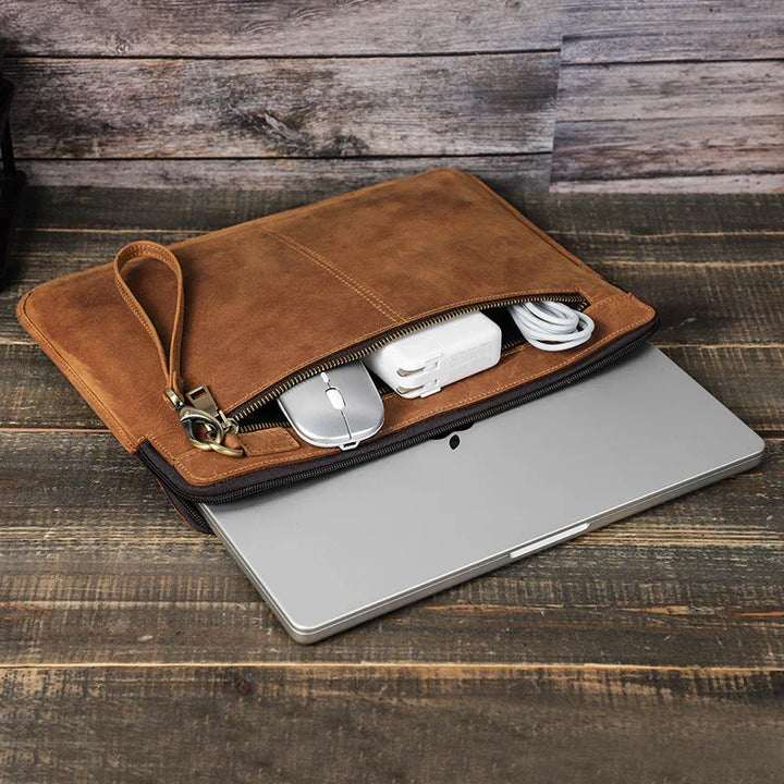 Brown leather laptop sleeve with zipper, holding a silver mouse, charger, and cable on wooden surface