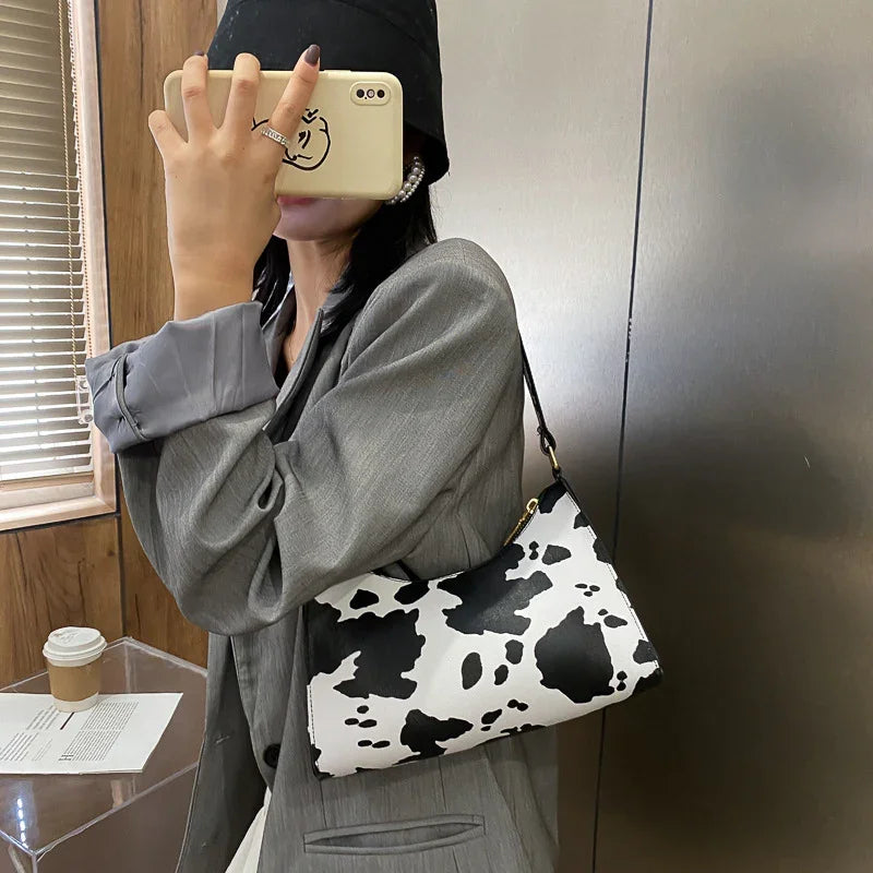 Woman wearing gray blazer and black bucket hat holding cow print shoulder bag, taking selfie indoors