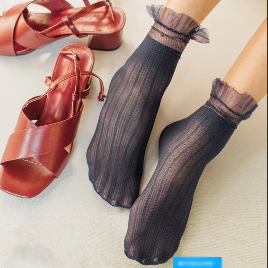 Feet wearing sheer black striped ankle socks with ruffled cuffs next to brown heeled sandals on beige surface