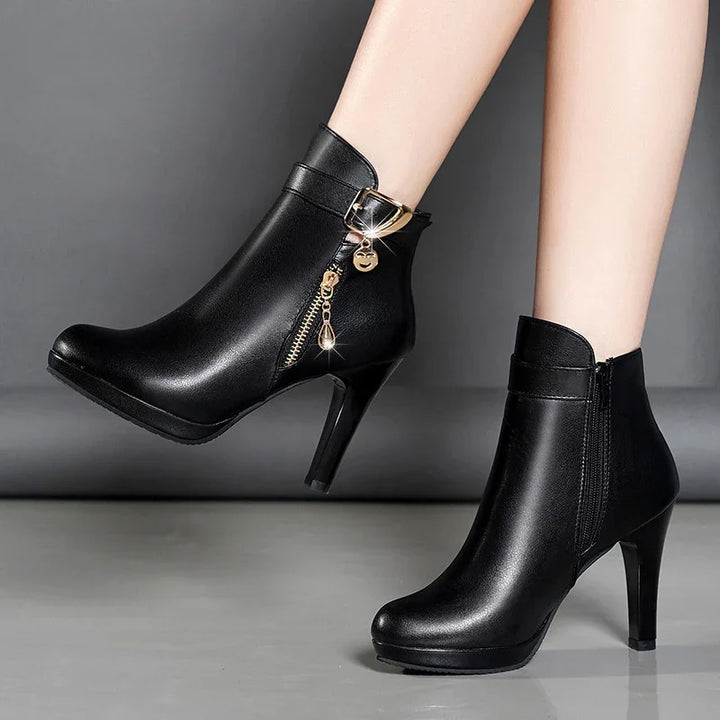 Black leather high-heeled ankle boots with gold zipper and charm detail on gray background