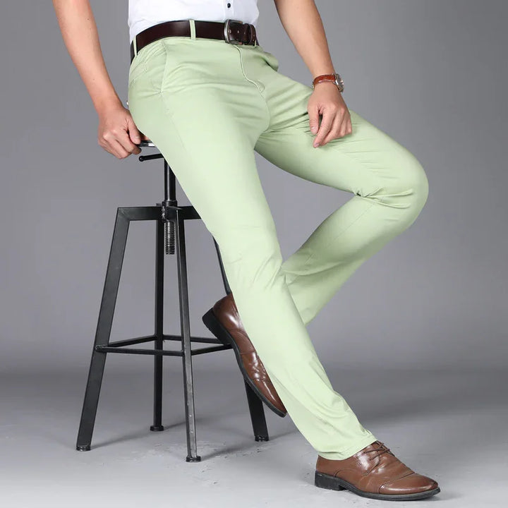 Man wearing light green slim-fit trousers with brown belt and brown dress shoes sitting on black metal stool