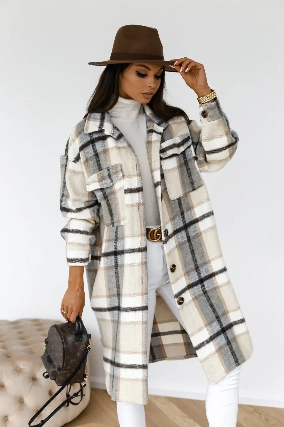 Woman wearing beige and gray plaid long coat, white turtleneck, brown wide-brim hat, holding designer handbag