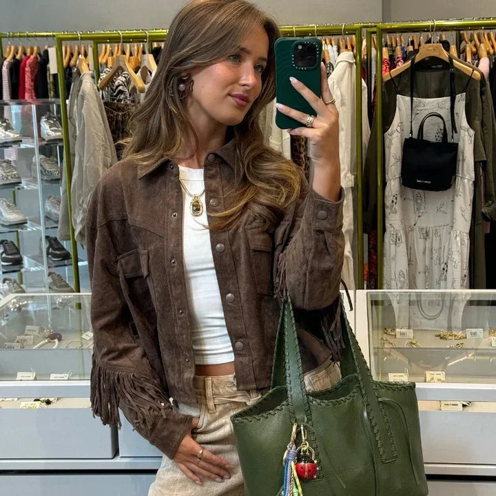 Woman in brown fringe jacket taking mirror selfie in boutique with green leather tote and jewelry displays