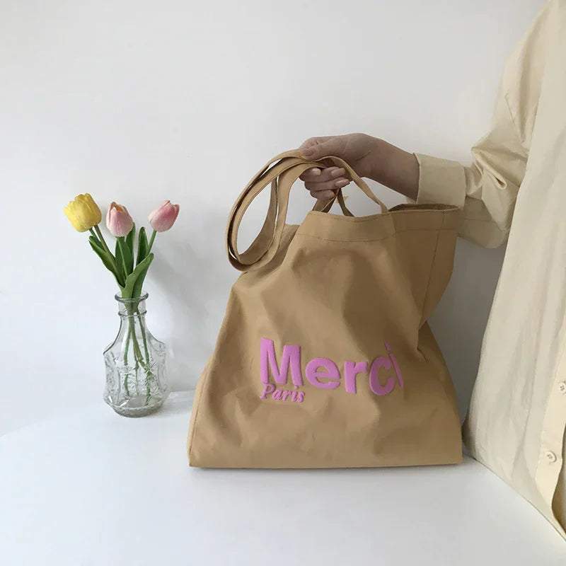 Beige canvas tote bag with pink Merci Paris text held by person in beige shirt next to clear vase with yellow and pink tulips