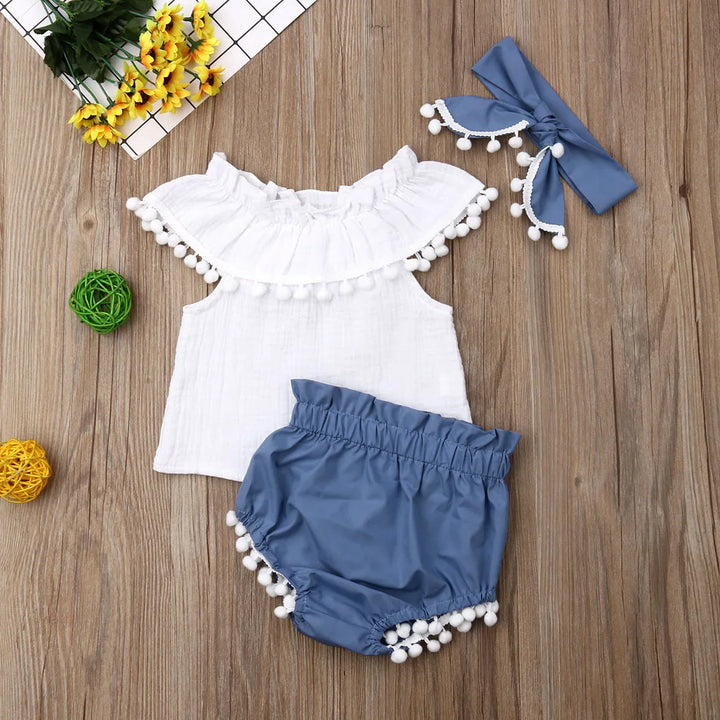 Infant summer outfit set with white ruffled top, blue shorts with pom-pom trim, and matching headband on wooden background