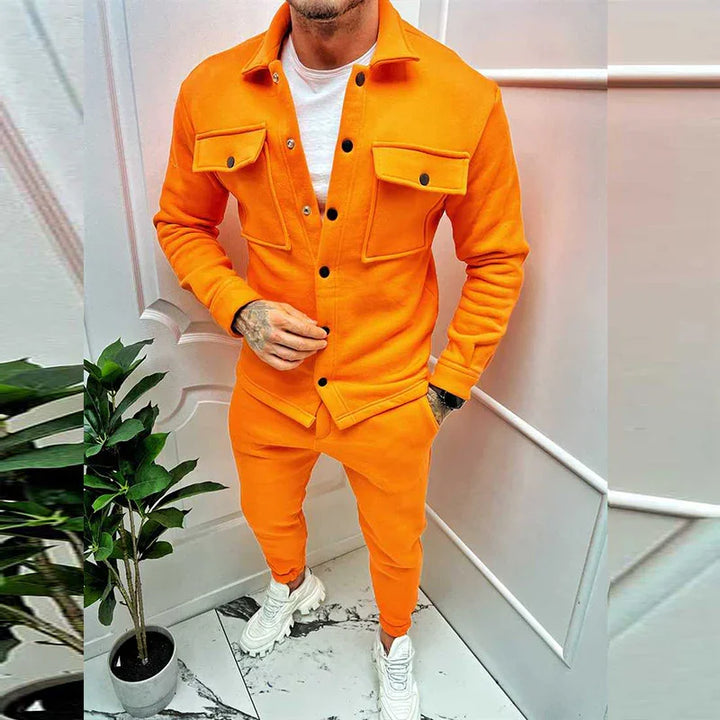 Man wearing bright orange jacket and jogger pants with white sneakers, standing indoors near plant