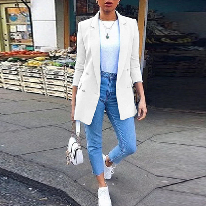 Woman in white blazer, blue jeans, and white sneakers walking on city sidewalk near fruit stand