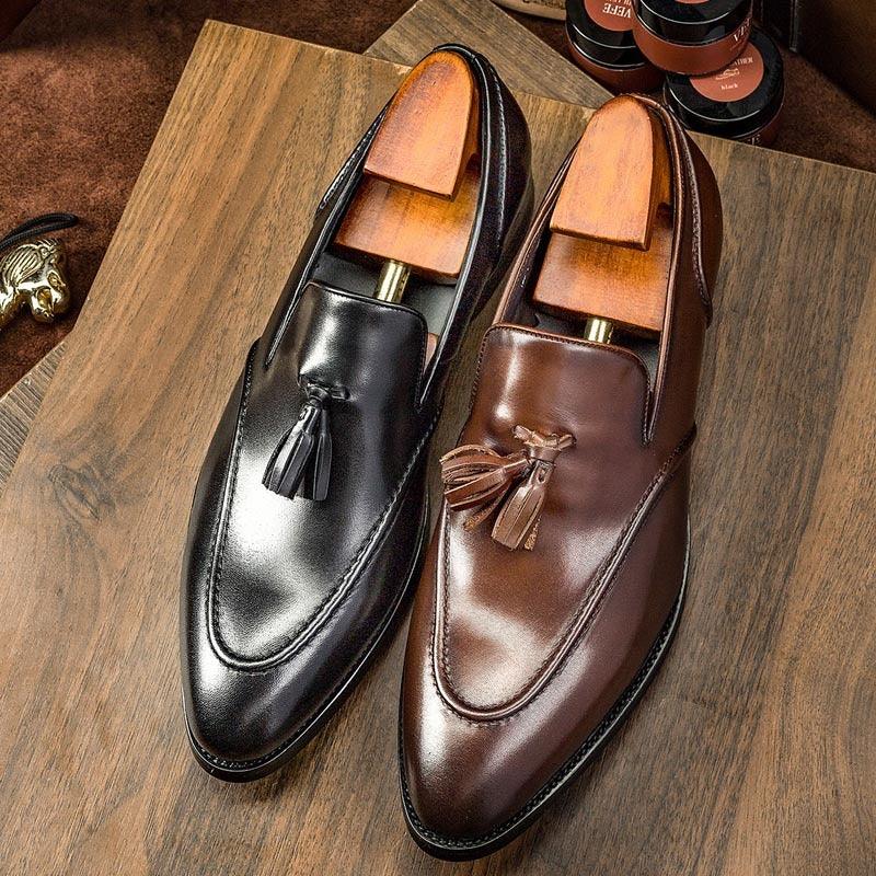 Pair of polished black and brown leather tassel loafers with wooden shoe trees on wooden surface
