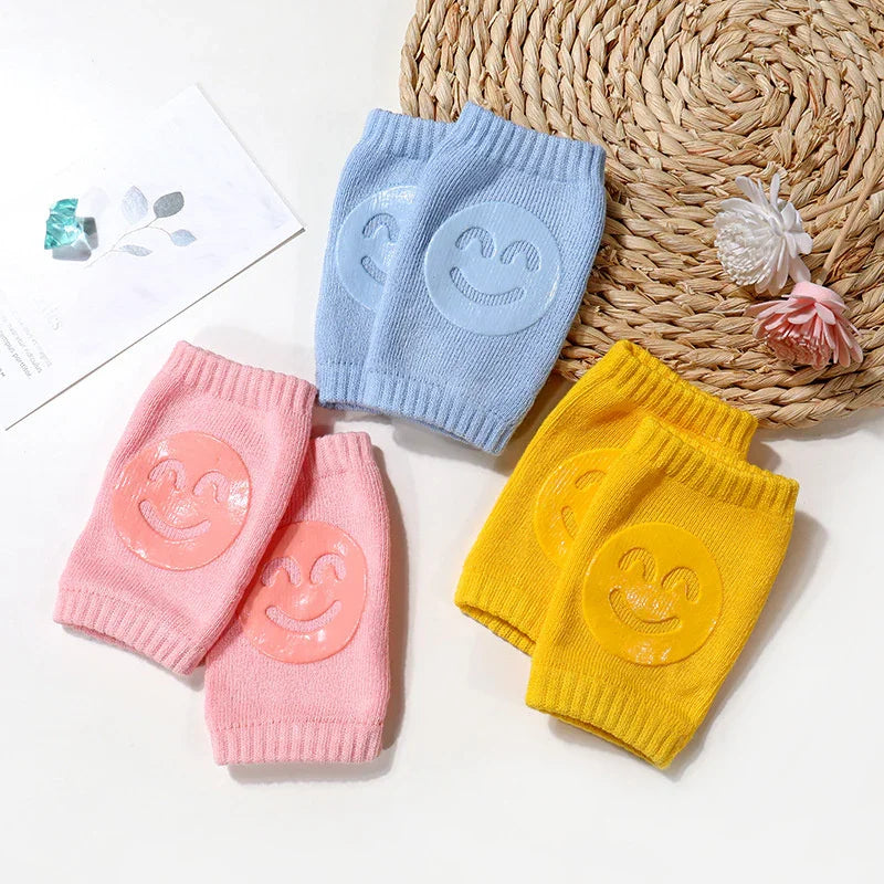 Colorful knitted baby knee pads in pink, blue, and yellow with smiling face prints on white background