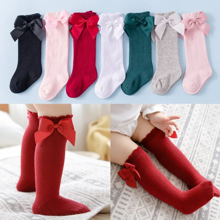 Colorful baby knee-high socks with large bows in black, pink, red, white, green, gray, and light pink, worn by baby