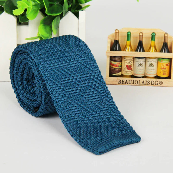 Rolled blue knitted necktie on white surface with green plants and miniature wine bottle crate in background