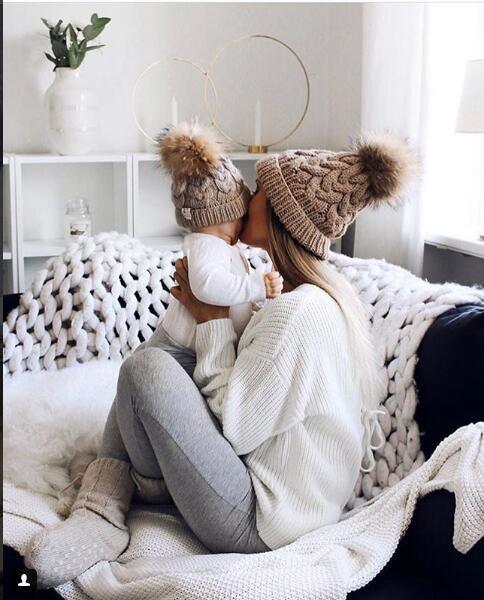 Mother and baby in matching beige knit hats with pom-poms cuddling on cozy white knit blanket in bright living room
