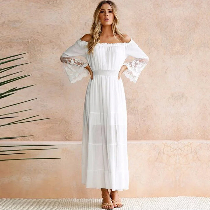 Woman in a white off-shoulder maxi dress with lace bell sleeves standing against a beige textured wall