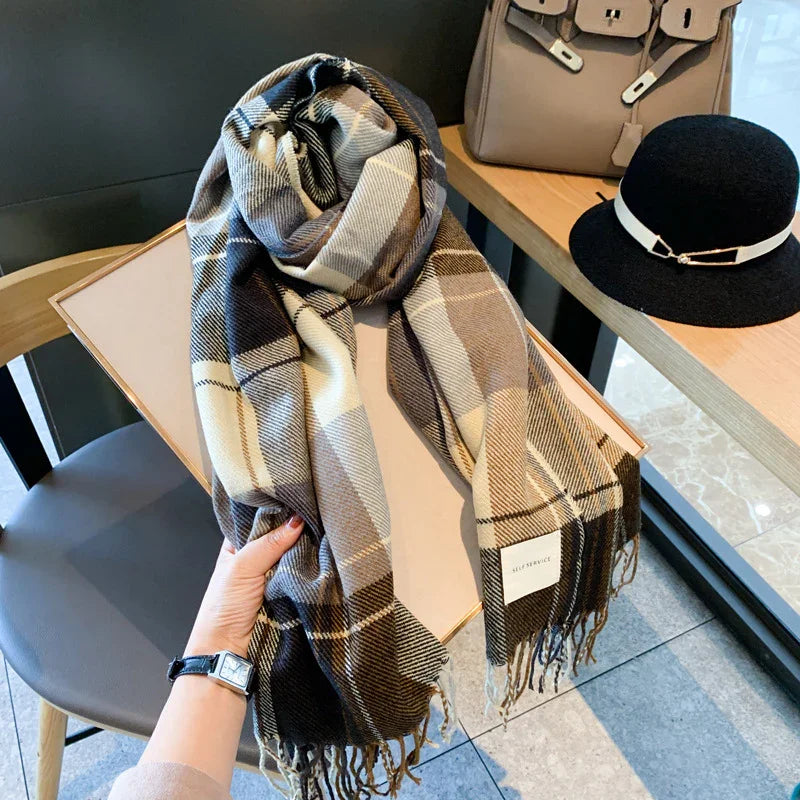 Hand holding a plaid wool scarf in brown and cream tones with fringe, next to black hat and beige handbag