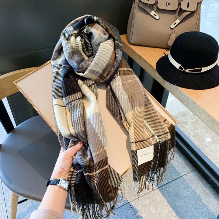 Hand holding a plaid wool scarf in brown and cream tones with fringe, next to black hat and beige handbag