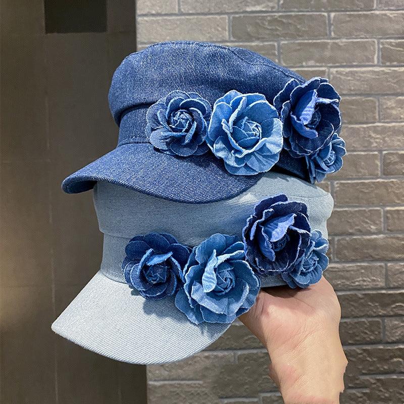 Hand holding two denim caps adorned with blue fabric flower decorations against a brick wall background