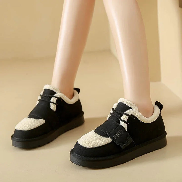 Black casual shoes with white shearling lining and strap detail, worn on bare feet indoors