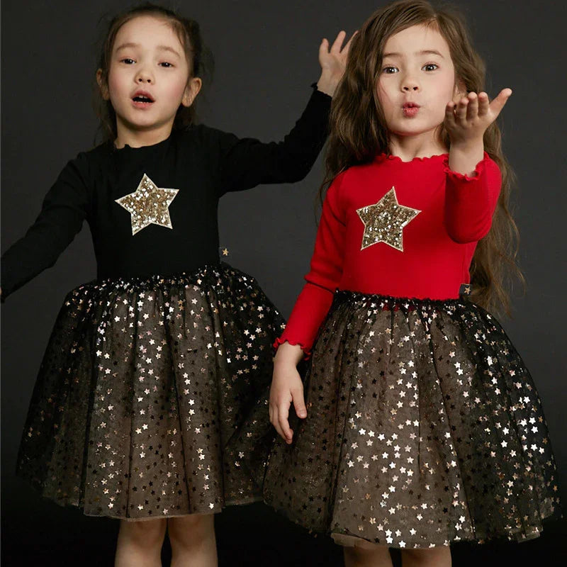 Two girls wearing glittery star dresses with black tulle skirts, one in black top and one in red top, posing against dark background
