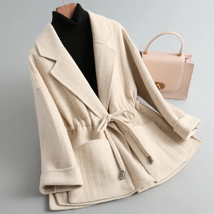 Beige wool blend women's coat with waist tie, wide lapels, and large front pockets, paired with black turtleneck and nude handbag