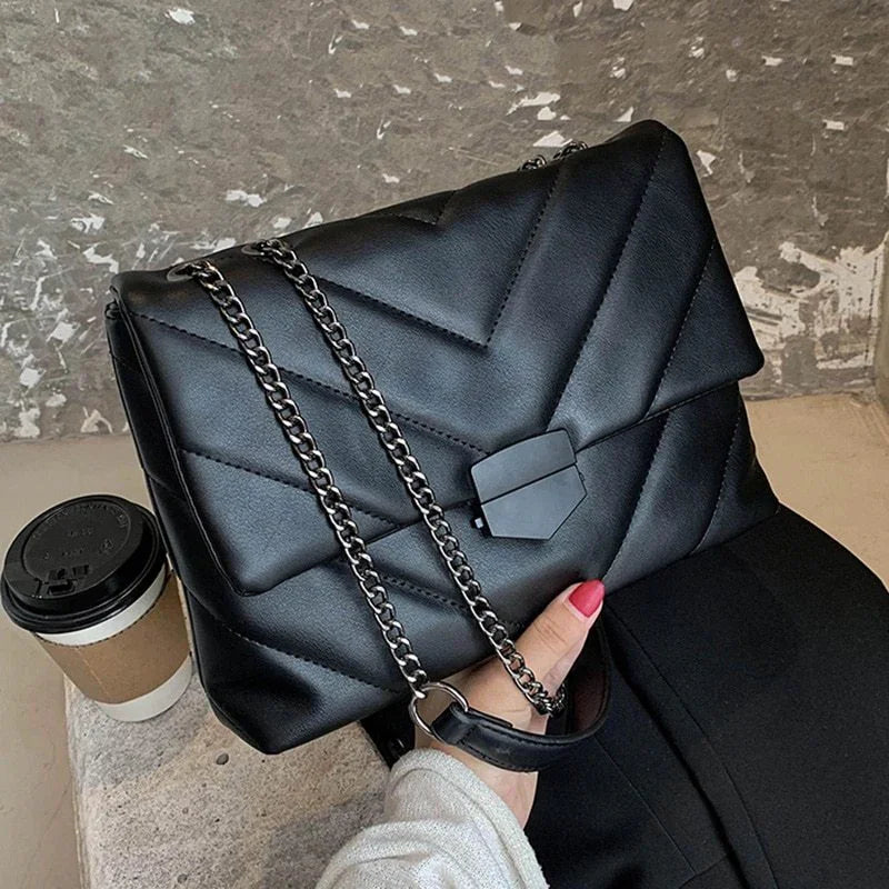 Black quilted leather handbag with silver chain strap held by hand, next to takeaway coffee cup