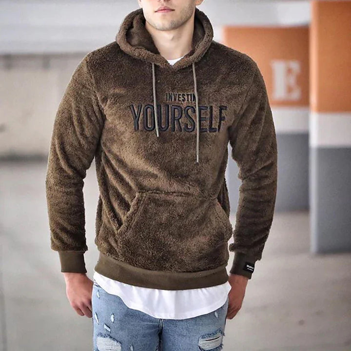 Man wearing brown fuzzy hooded sweatshirt with 'Invest In Yourself' text in urban setting