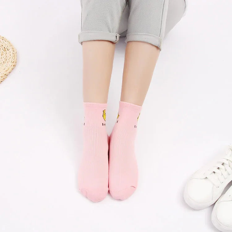 Pair of feet wearing pink ankle socks with yellow cartoon prints, light grey cuffed pants, white sneakers nearby