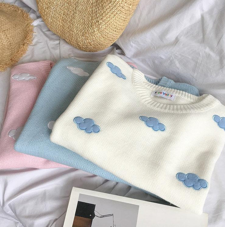 Folded pastel sweaters with embroidered cloud designs on white fabric background with straw hats