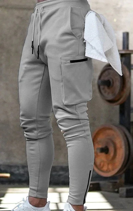 Man wearing light gray cargo jogger pants with side pockets and zippers, white sneakers, gym setting background