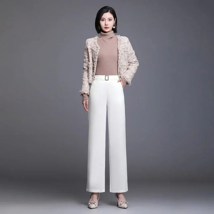 Woman in beige turtleneck sweater, textured jacket, white wide-leg pants, and beige heels against gray background