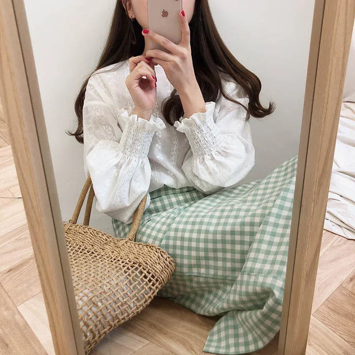 Woman taking mirror selfie wearing white long-sleeve blouse and green gingham skirt with woven straw bag