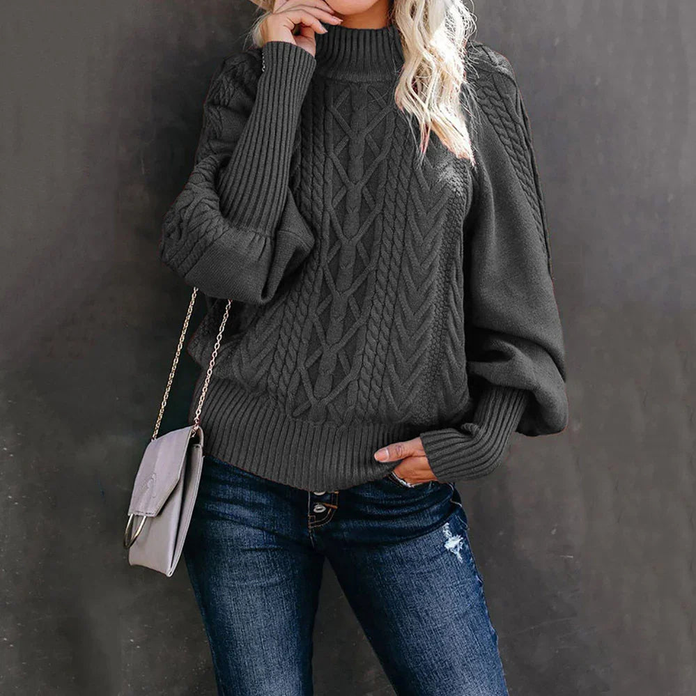 Woman wearing dark gray cable knit sweater with high neck and blue distressed jeans, carrying gray crossbody bag