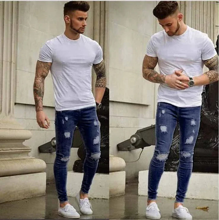Man with tattooed arms wearing fitted white t-shirt, ripped skinny jeans, and white sneakers in urban setting