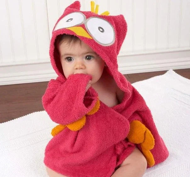 Baby wearing red owl hooded towel with big eyes and orange wings sitting on white blanket indoors