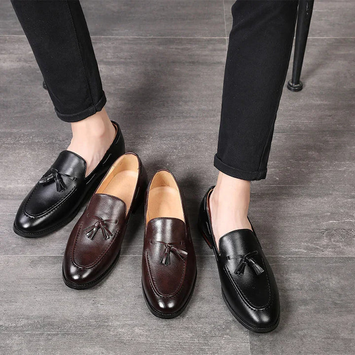 Men's tassel loafers in black and brown leather on gray floor with model wearing black pants