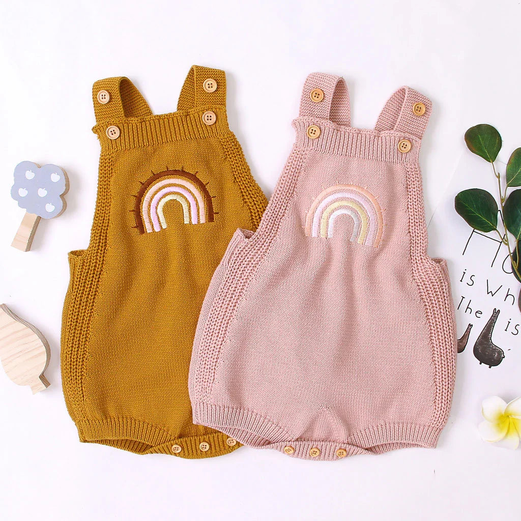 Two knitted baby rompers with rainbow embroidery, one mustard yellow and one light pink, with wooden buttons