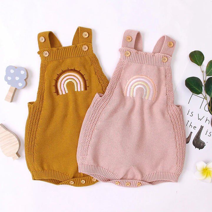 Two knitted baby rompers with rainbow embroidery, one mustard yellow and one light pink, with wooden buttons