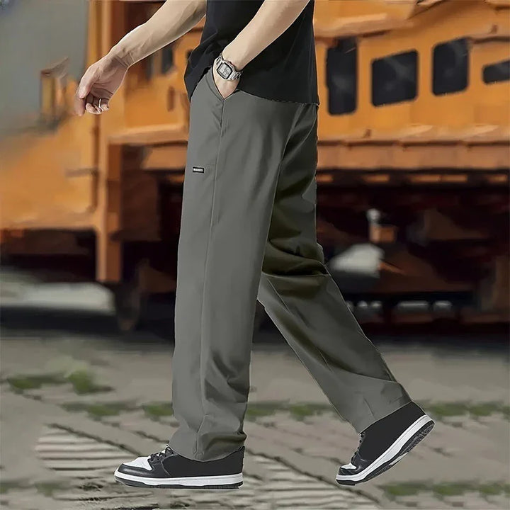 Person wearing dark gray casual pants and black sneakers walking outdoors with orange industrial background