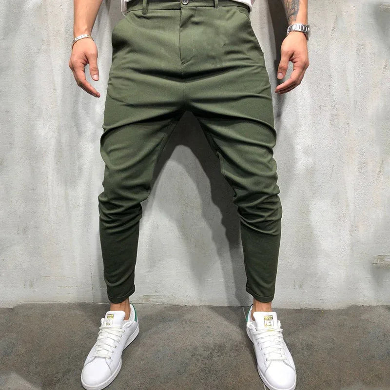 Man wearing olive green tapered pants and white sneakers standing against gray concrete wall