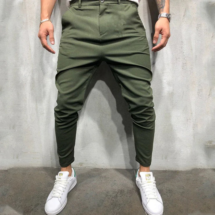 Man wearing olive green tapered pants and white sneakers standing against gray concrete wall