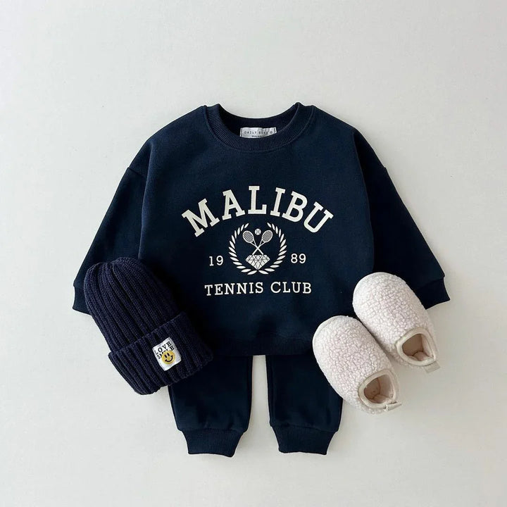 Navy blue toddler outfit with Malibu Tennis Club sweatshirt, matching pants, knit beanie, and white fuzzy slippers