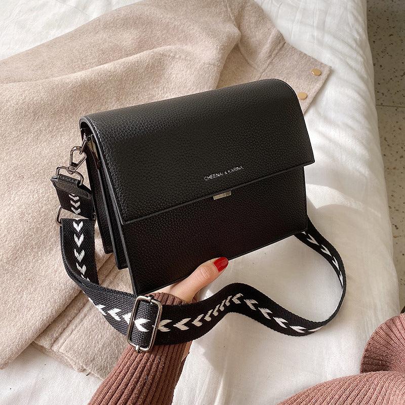 Black textured leather crossbody bag with white patterned strap on beige fabric background