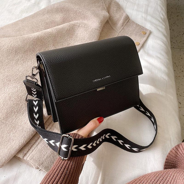 Black textured leather crossbody bag with white patterned strap on beige fabric background