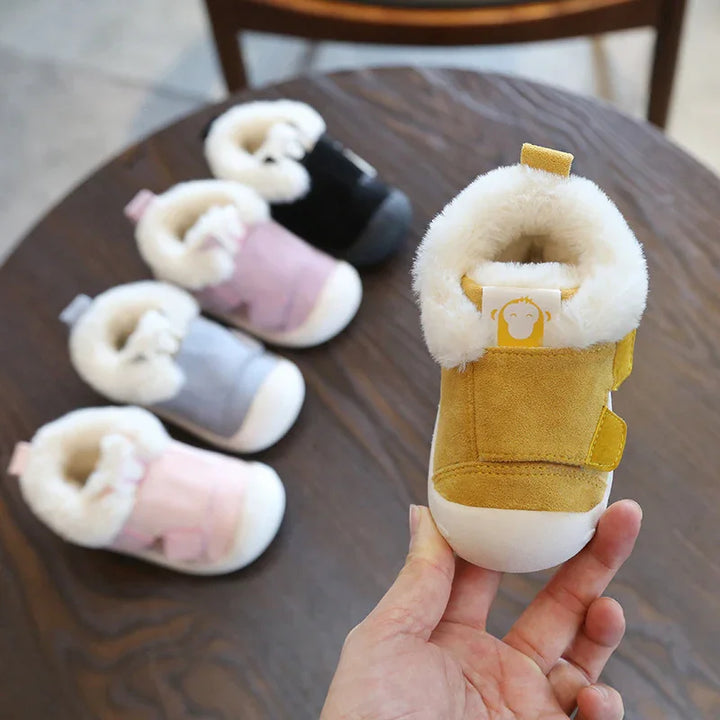 Hand holding yellow baby bootie with fur lining, five other fur-lined baby shoes in pink, gray, black on wooden surface