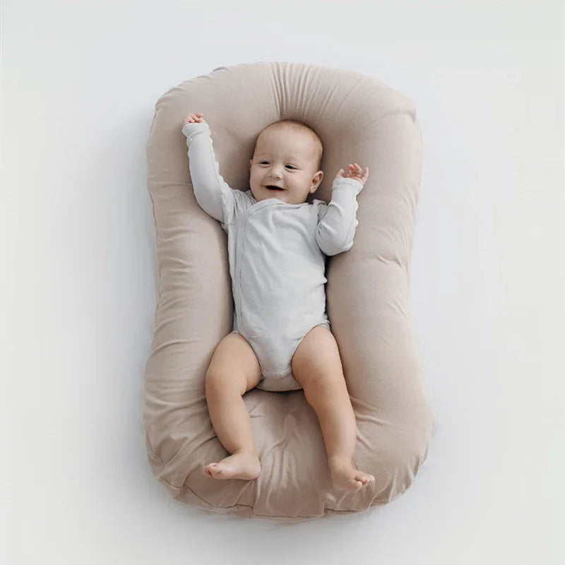 Happy baby in light grey onesie lying on beige soft cushioned baby lounger on white background