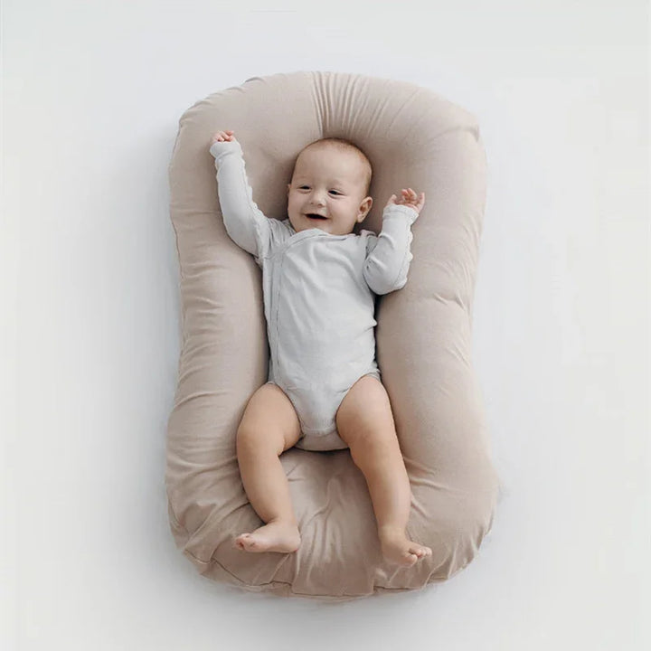 Happy baby in light grey onesie lying on beige soft cushioned baby lounger on white background