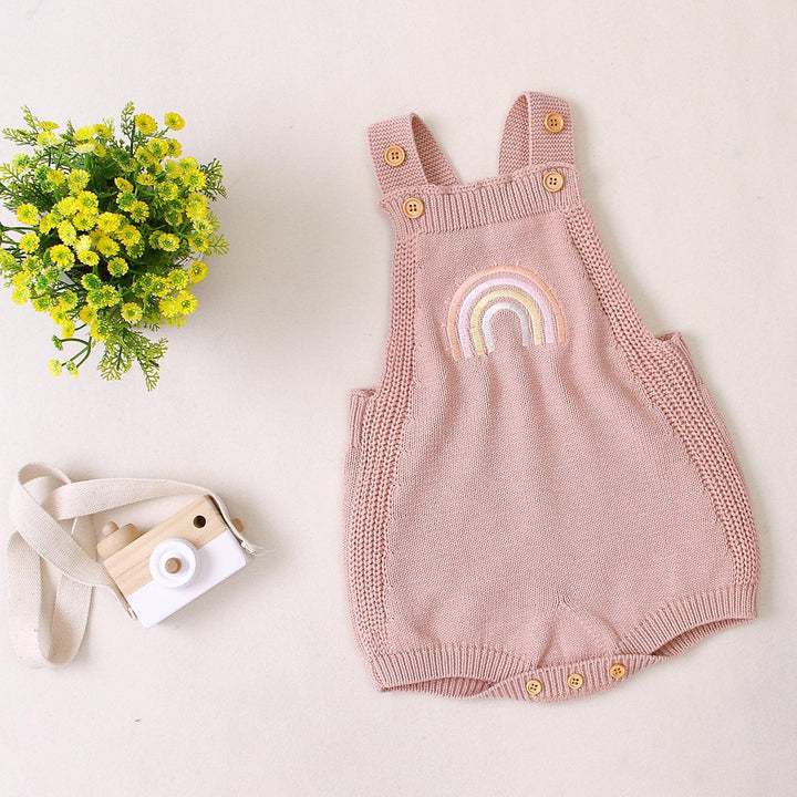 Pink knitted baby romper with rainbow embroidery, wooden buttons, toy camera, and yellow flowers