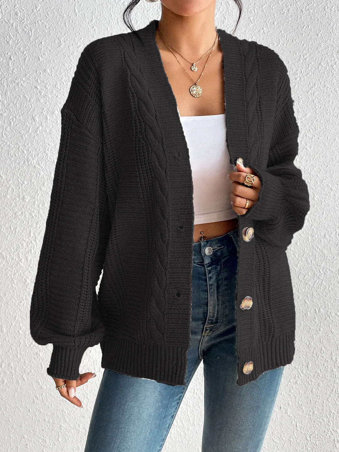 Woman wearing black cable knit button-up cardigan, white crop top, blue jeans, and layered gold necklaces