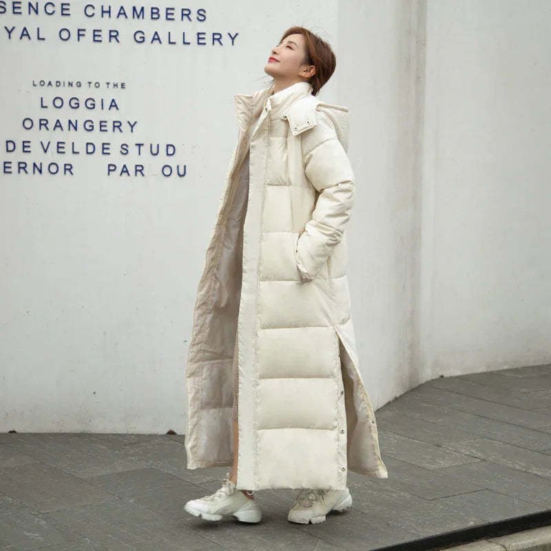 Woman wearing a long cream puffer coat and white sneakers standing against white wall with black text